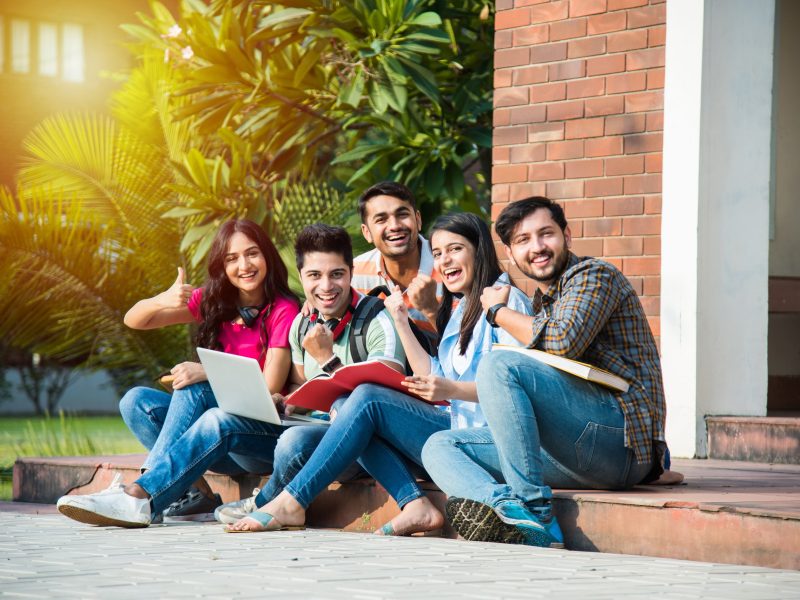Young Asian Indian college students reading books, studying on laptop, preparing for exam or working on group project while sitting on grass, staircase or steps of college campus